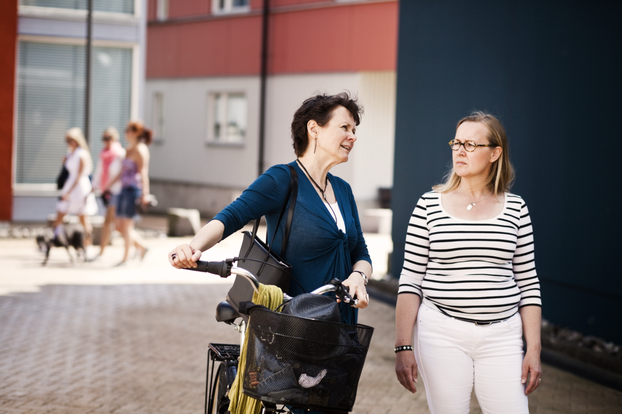 Zwei erwachsene Frauen unterhalten sich auf der Straße. Eine Frau schiebt ein Fahrrad.