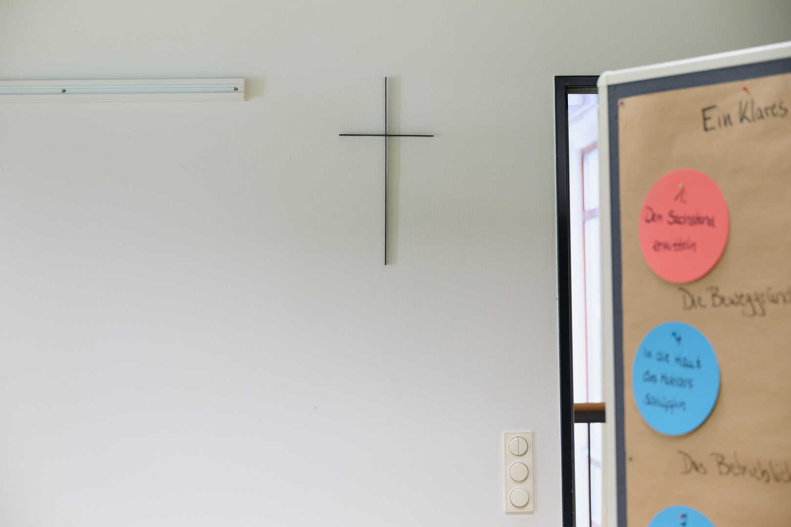 Kreuz hängt an einer weißen Wand vor einer beklebten Pinnwand